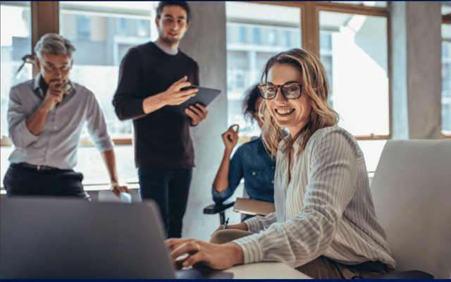 Female sitting colleague working on computer and others behind her smiling