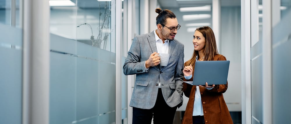 Man and woman walking in office hallway with coffee and laptop